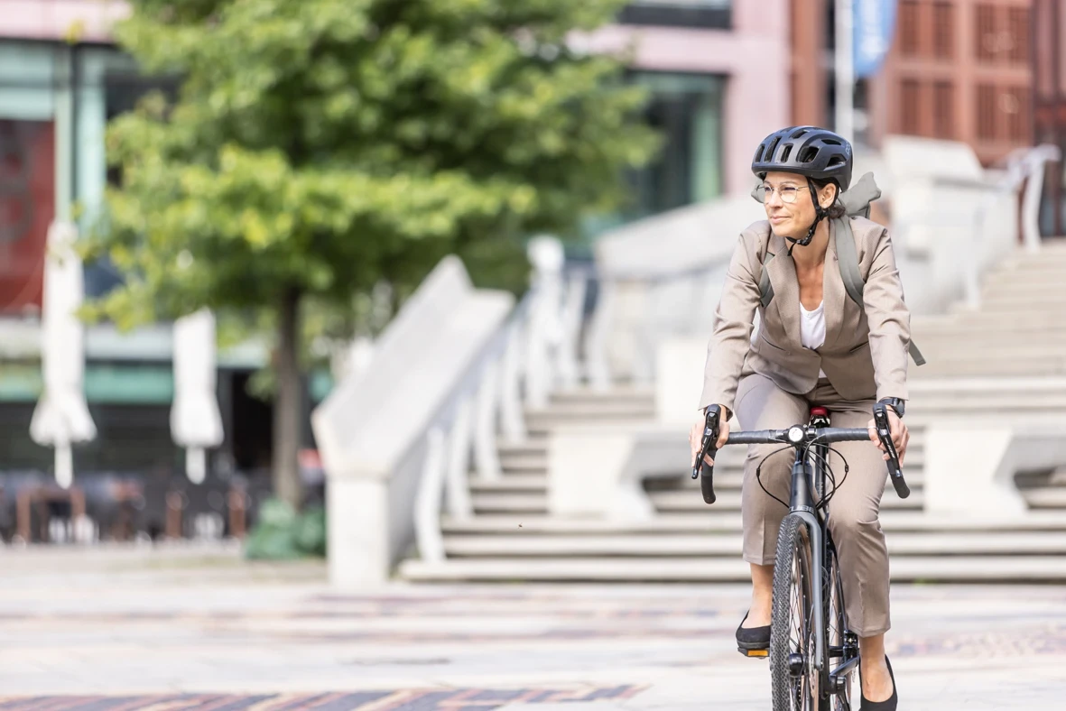 Frau in Gesch&auml;ftskleidung f&auml;hrt mit Helm auf einem Fahrrad durch eine urbane Platzanlage.