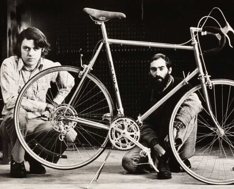 Zwei M&auml;nner posieren mit Fahrr&auml;dern in einer Werkstatt.