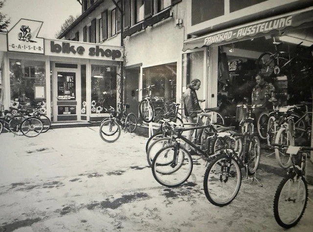 Historische Schwarz-Wei&szlig;-Aufnahme einer Fahrradwerkstatt mit ausgestellten R&auml;dern vor dem Gesch&auml;ft.