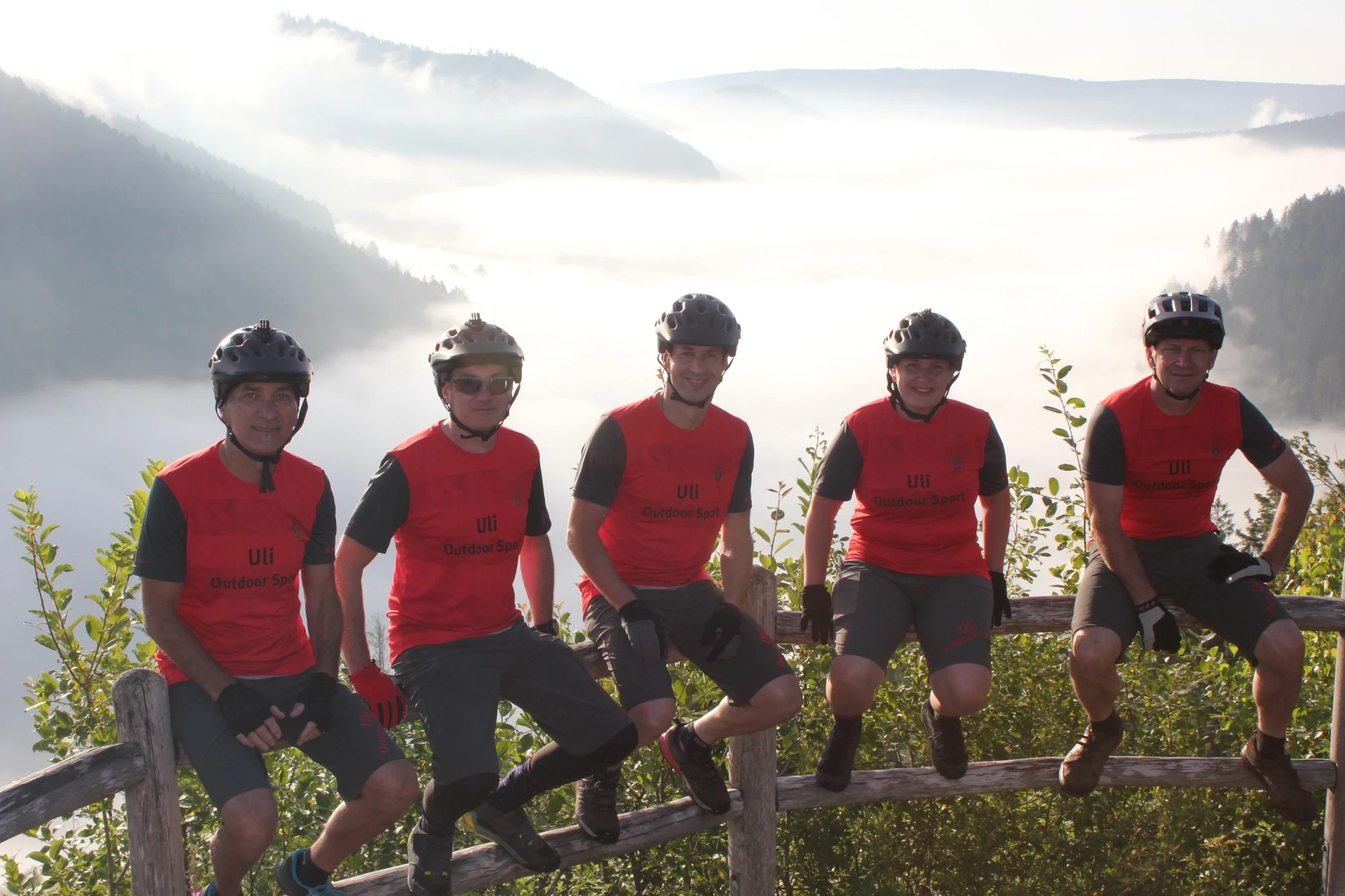 F&uuml;nf Radfahrer in roten Trikots stehen auf einem Aussichtspunkt mit Bergblick und Nebel.