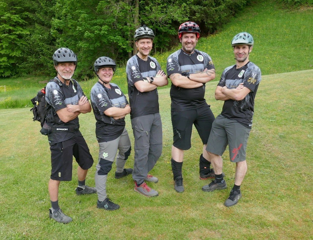 Mann mit Fahrradhelm und schwarzem Trikot steht l&auml;chelnd vor gr&uuml;ner Wiese und Wald.