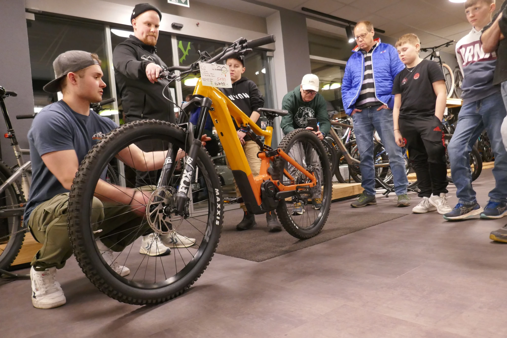 Mann pr&auml;sentiert oranges Mountainbike einer Gruppe von Besuchern in einer Werkstatt.