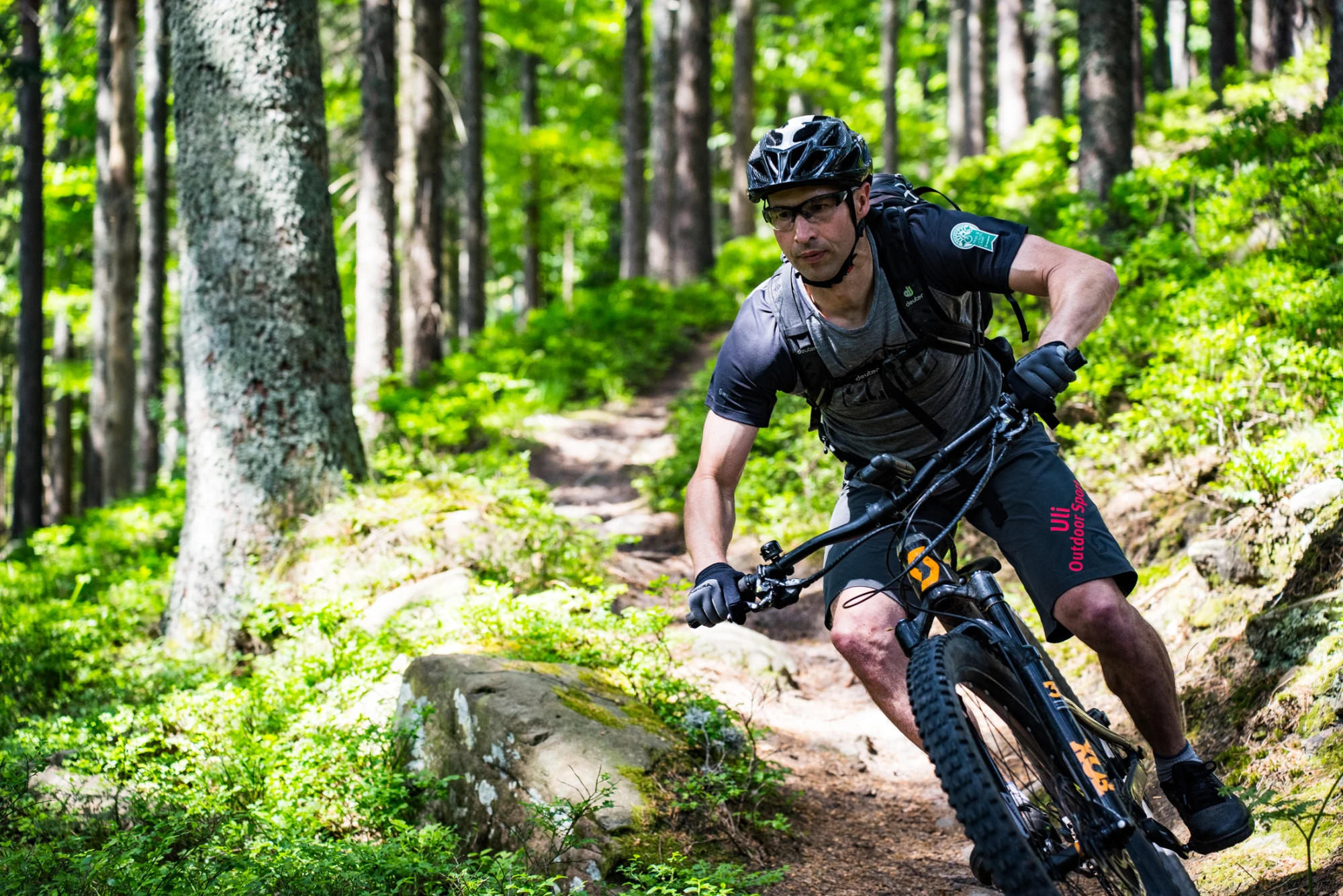 Drei Radfahrer mit Fahrr&auml;dern stehen an einer Aussichtsplattform mit Berglandschaft im Hintergrund.