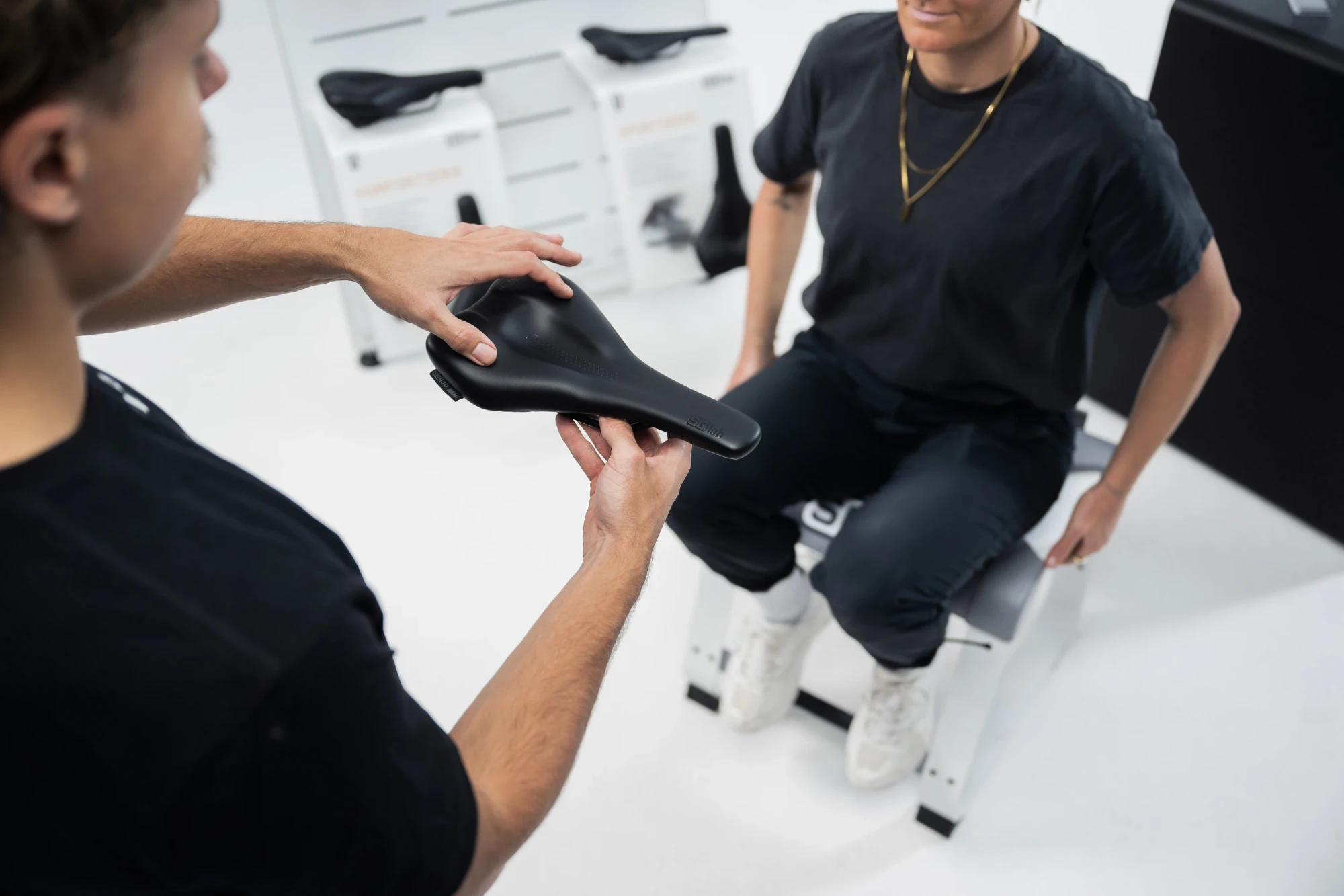 Zwei Personen begutachten einen schwarzen Fahrradsattel in einer Fahrradwerkstatt.