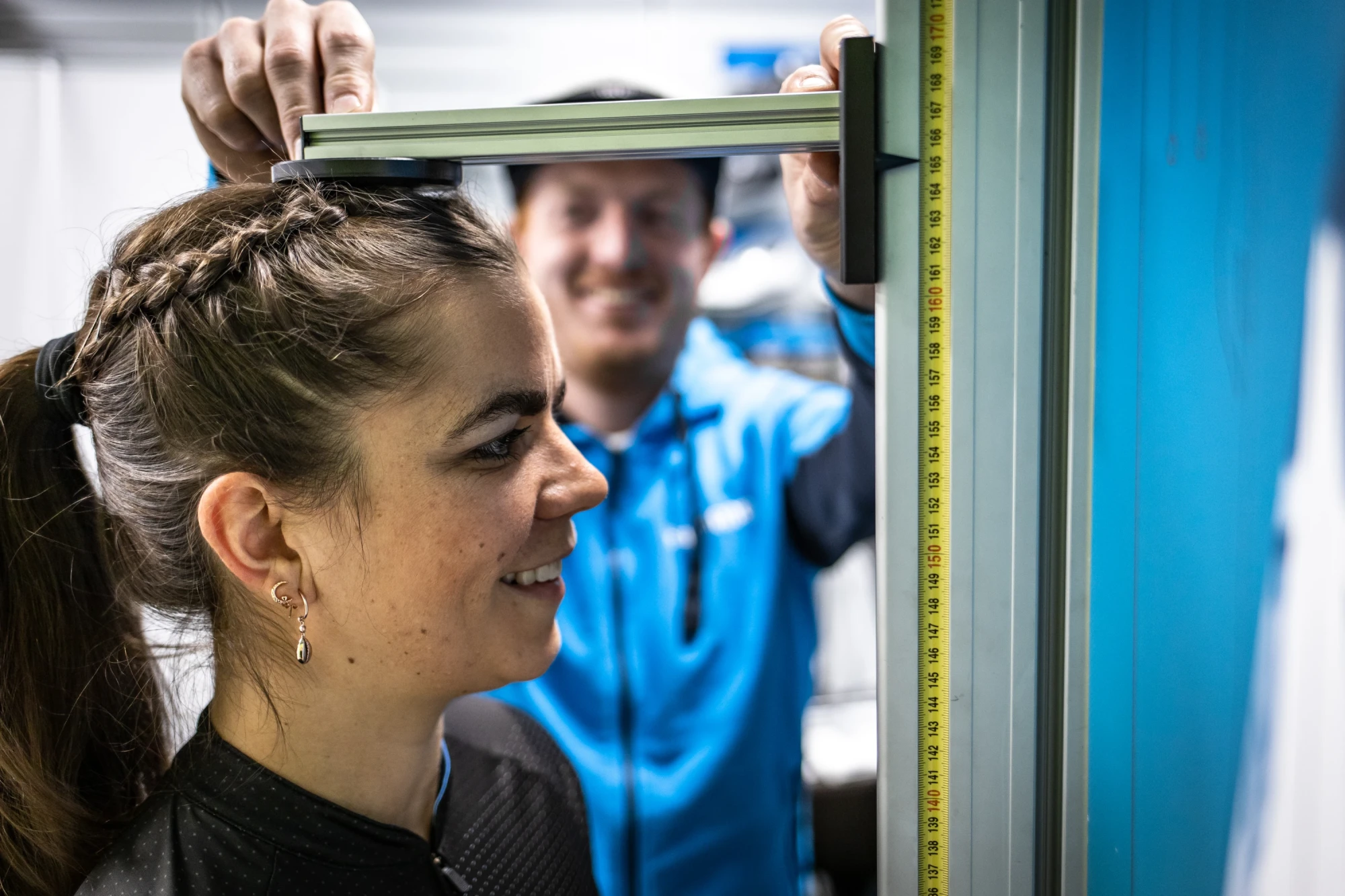 Eine Frau misst mit einem Stahlma&szlig;stab die H&ouml;he eines Fensterrahmens, w&auml;hrend ein Mann in blauer Arbeitskleidung im Hintergrund zuschaut.