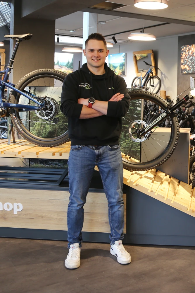 Junger Mann mit verschr&auml;nkten Armen l&auml;chelt in die Kamera, tr&auml;gt schwarzes Gaiser-Shirt in Fahrradwerkstatt.