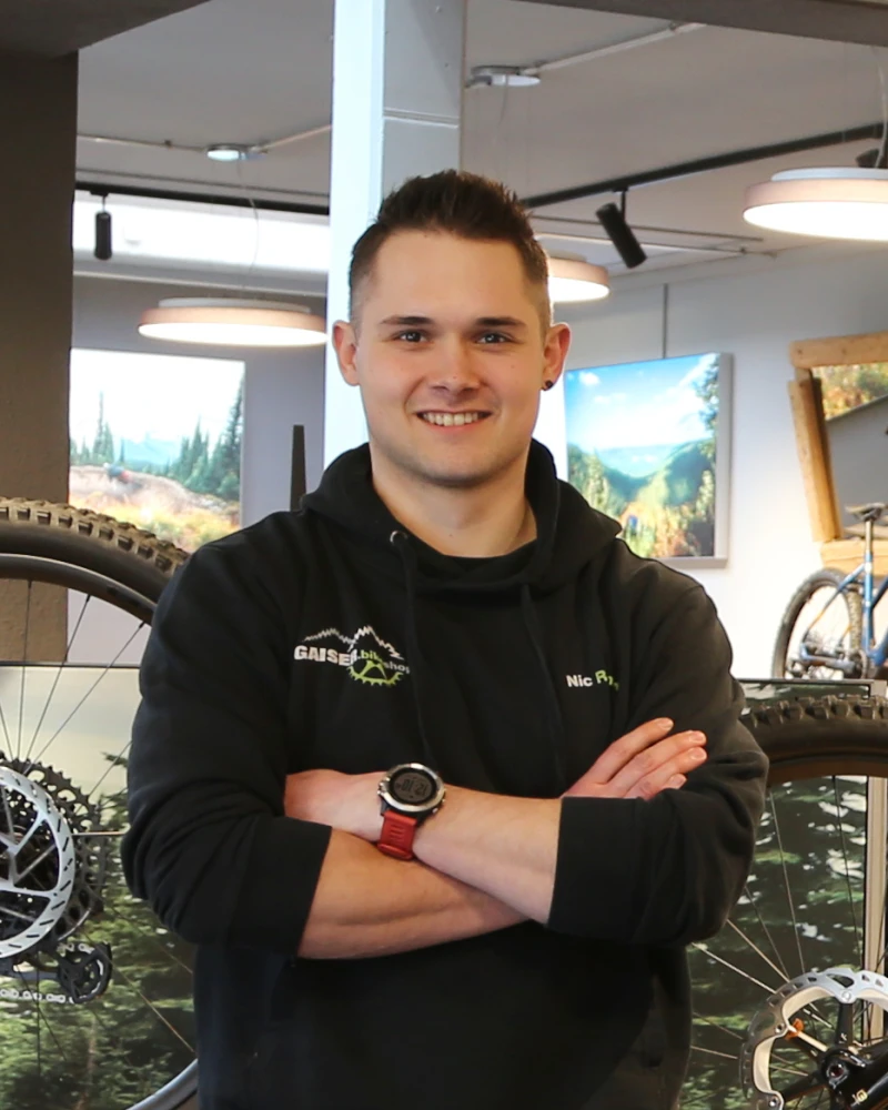 Junger Mann mit verschr&auml;nkten Armen l&auml;chelt in die Kamera, tr&auml;gt schwarzes Gaiser-Shirt in Fahrradwerkstatt.