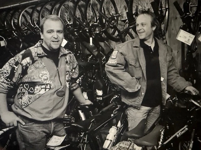 Zwei M&auml;nner stehen in einer Fahrradwerkstatt zwischen aufgeh&auml;ngten Fahrr&auml;dern.