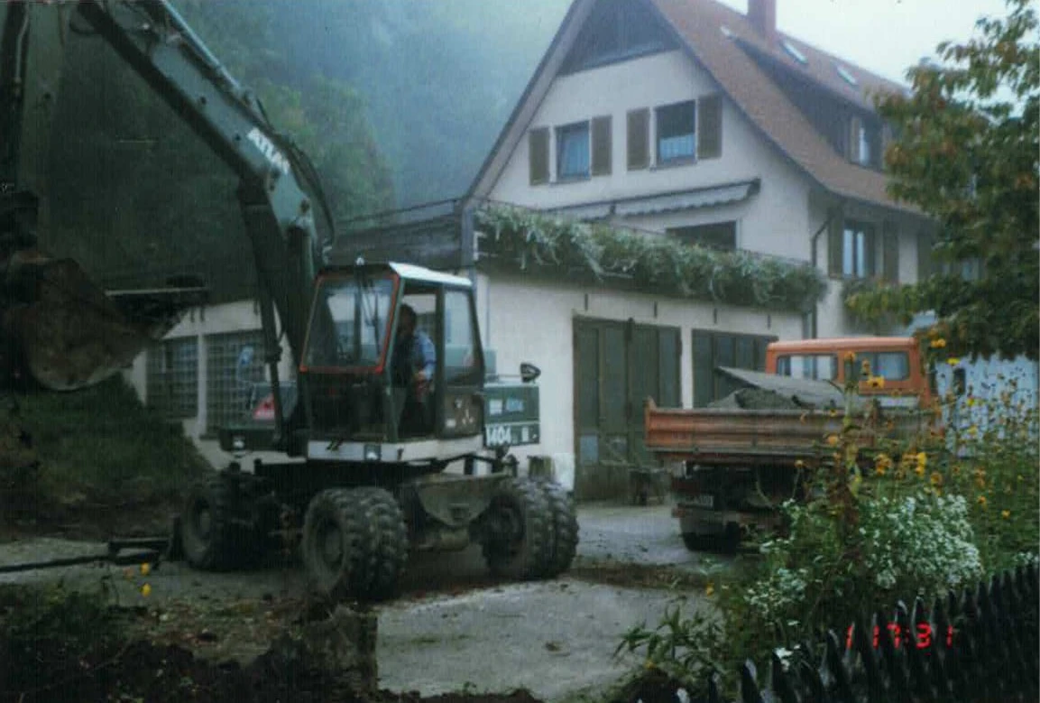 Radlader und Holzanh&auml;nger vor wei&szlig;em Fachwerkhaus mit Garten.